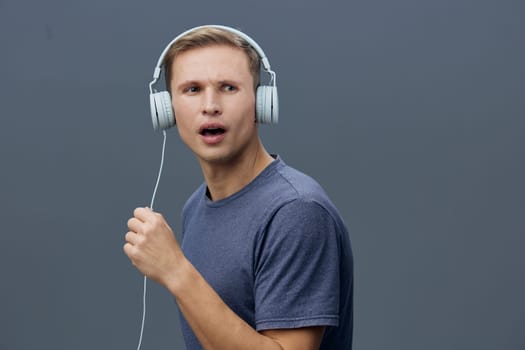 Young man with short hair and fair skin enjoying music through big headphones, expressing surprise or confusion, casual outfit, gray background, studio shot, emphasizing entertainment or sound experience.