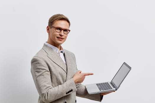 Confident young man wearing formal suit and glasses holding and pointing to open laptop on a plain white background. Business, technology, professional, presentation, communication, success concepts.