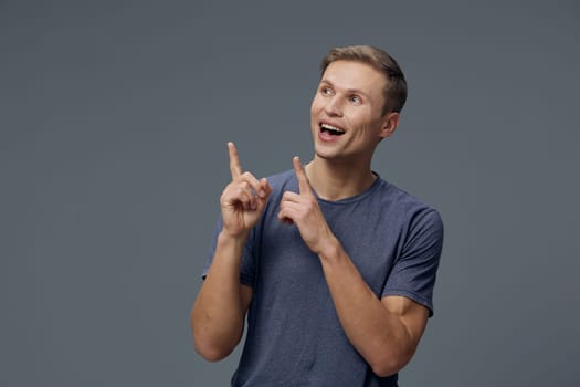 smiling young man in casual blue t-shirt point fingers upward with excited face on studio gray background. cheerful man express positive emotions, digital detox and mental health with gadgets concept. casual lifestyle and technology.