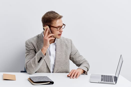 Young professional man with glasses, dressed in a gray blazer and white shirt, sitting at a clean desk using a laptop, talking on a smartphone, and writing notes, isolated on white background. Business, communication, work life