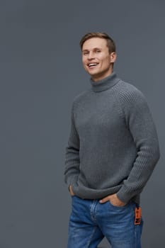 Smiling man dressed in a cozy gray turtleneck sweater and blue jeans stands confidently against a plain gray background, expressing happiness and relaxed style with a warm and inviting look. Concept: casual elegance