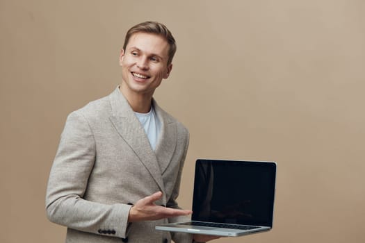 Cheerful young man with bright smile presenting open laptop computer while standing against plain beige backdrop. Experienced in freelance work, technology enthusiast, casual style, modern professional, youthful energy, digital tools, positive attitude. Concept: modern remote work