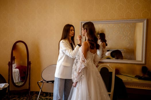A stylist or makeup artist prepares a bride for a wedding indoors. The backdrop is a warm-toned hotel interior with mirrors