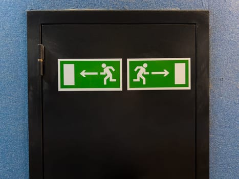 Two emergency exit signs with running figure and arrows pointing left and right placed on black door panel against blue wall, providing clear direction for safe evacuation in case of emergency