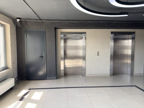Modern building interior with two stainless steel elevators, dark utility door, surveillance camera and geometric ceiling light, illustrating clean minimalist hallway in residential or office space
