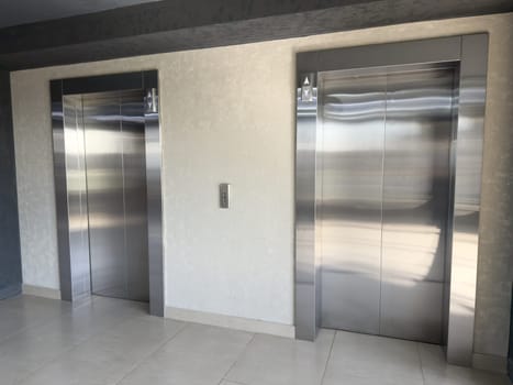 Closeup of two modern stainless steel elevator doors in light tiled hallway with textured wall, reflecting ambient light and emphasizing clean lines, symmetry and minimalism in architectural space