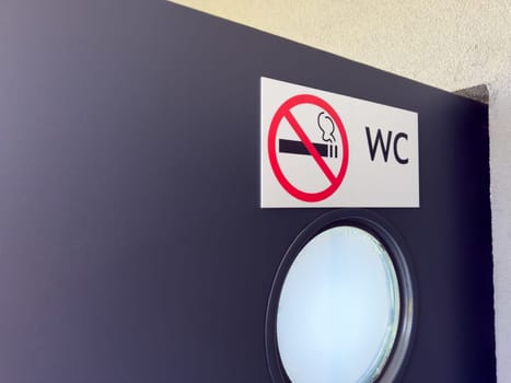 Closeup of WC door with round frosted window and no smoking sign on white label, showing clear visual message for public restroom users in clean and modern architectural setting