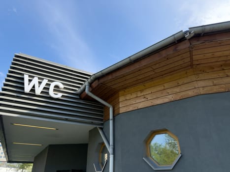 Modern public restroom building with large WC letters on metal facade and wooden roof detail under blue sky, combining practical function with clean architectural design in outdoor urban space