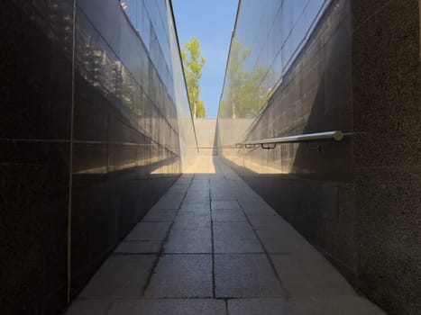 Urban ramp with polished granite walls and textured stone floor leading to daylight, showcasing contrast between hard materials and reflective surfaces in clean symmetrical architectural layout