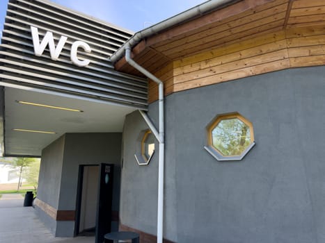 Contemporary public restroom with large WC sign, open door entrance, wooden roof detail and octagonal windows, combining practical accessibility with clean architectural elements in park environment