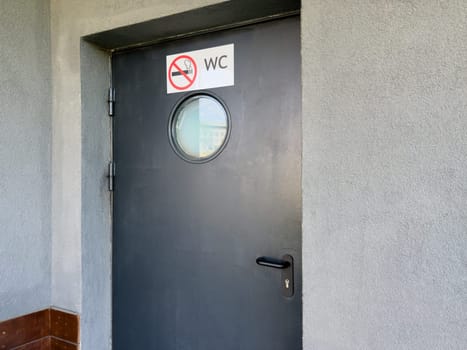 Black door with round frosted window and no smoking WC sign mounted on rough gray wall, marking entrance to public restroom with emphasis on clarity, hygiene and behavioral regulation in urban setting