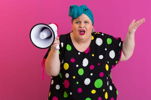 Confident woman in a colorful polka dot outfit and retro headwrap yelling into a megaphone, expressing strong emotion and attitude against a bold pink background.