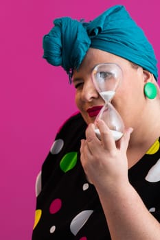 Portrait of a confident woman with a retro headwrap and colorful outfit, holding an hourglass in front of her face as a symbol of time and focus, with a vibrant pink background.