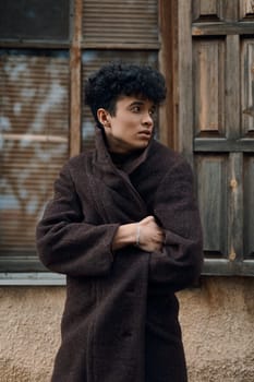 Fashionable young man with curly hair wearing a brown coat, standing in front of an old building with a window.