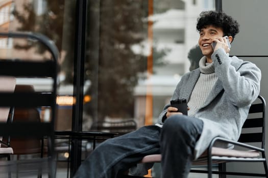 Man sitting on chair by glass window, talking on cell phone with coffee cup in hand. fashionable and stylish concept.