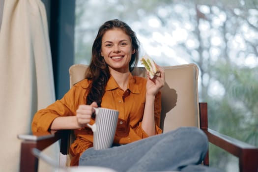 A cheerful young woman in an orange shirt holds a sandwich and a cup of tea, radiating happiness The soft background suggests a cozy, homely setting perfect for relaxation