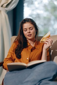 A young woman with long hair, dressed in an orange shirt and blue jeans, seated comfortably, enjoying a sandwich and engrossed in a book Bright and inviting indoor ambiance