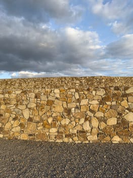 A sturdy stone wall stands against a backdrop of fluffy clouds in an expansive landscape. The sun sets, casting warm light on the textured stones and creating a serene atmosphere.