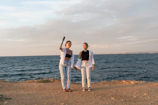 Two women stand on a beach with a body of water in the background, one holding a selfie stick. They are both wearing white jackets.