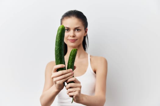 A young woman in a white tank top is cheerfully holding two fresh green cucumbers against a bright white background This image emphasizes health, vitality, and the importance of fresh produce in a healthy diet