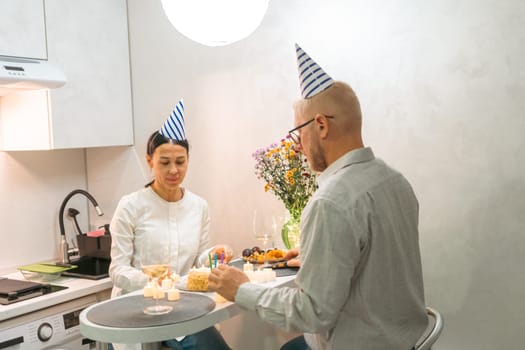 Here's a caption fitting your specifications: A bald man and a woman, both wearing party hats, sit at a kitchen table, sharing a meal. They appear to be celebrating a birthday.