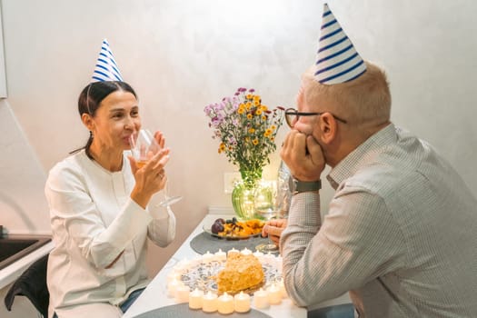 Here's a caption that fits the requirements: A happy couple celebrates a birthday. They wear party hats, sit at a table with cake and flowers. She claps; he smiles. An intimate birthday scene.