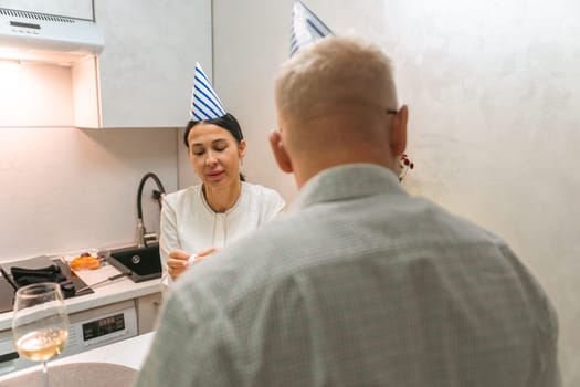 Here's a caption under 150 characters describing the image: A woman in a white top and a bald man, wearing a party hat, converse in a small kitchen. A celebratory scene.