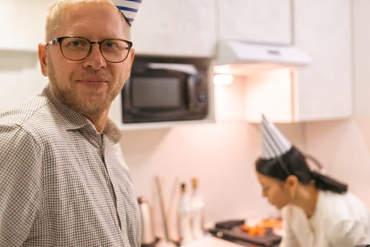 Here's a caption describing the image: A middle-aged man with glasses stands in a kitchen, slightly out of focus. Behind him, a person wearing a chef's hat prepares food at a counter. A microwave is visible on the wall.