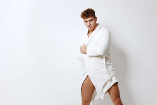 Stylish young man wearing a soft white bathrobe, standing confidently with a serious expression. Minimalist background enhances the focus on fashion and grooming.