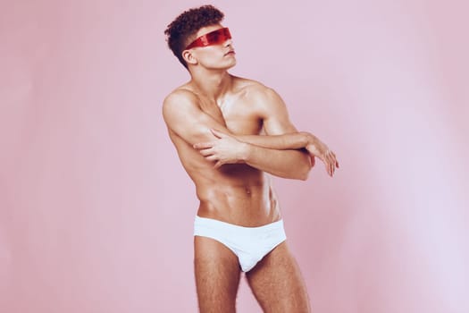 Stylish young man confidently posing in white swimwear and red sunglasses, set against a vibrant pink background. Emphasizing modern fashion and self-expression.