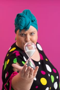 Portrait of a confident woman with a retro headwrap and colorful outfit, holding an hourglass in front of her face as a symbol of time and focus, with a vibrant pink background.