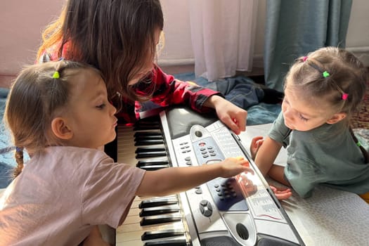 Teenage boy sits on the floor with his twin toddler sisters, showing them how to play a synthesizer at home. Happy family time and early music education concept