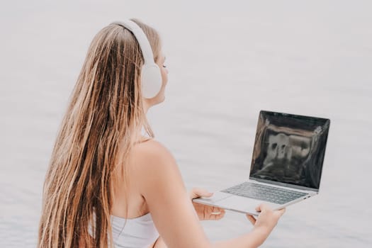 A woman is sitting by the water with a laptop in her lap. She is wearing headphones and she is listening to music