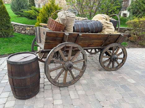 A rustic wooden cart stands in a garden, filled with barrels and burlap sacks, surrounded by lush greenery and stone landscaping. This scene evokes a nostalgic farming atmosphere.