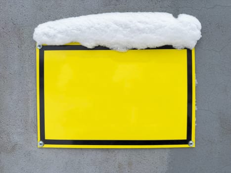 A vibrant yellow sign is attached to a gray wall, partially hidden under a layer of snow. The contrasting colors create a striking visual effect in a winter environment.