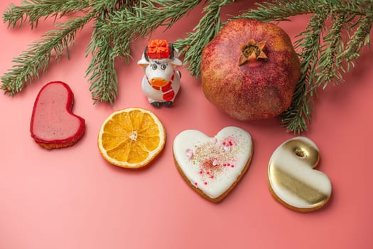 Colorful cookies shaped like hearts, dried orange slices, and a pomegranate are arranged with a small cow figurine and pine branches on a pink background, creating a joyful holiday display.