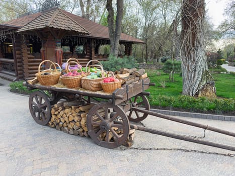 A wooden cart stands prominently, showcasing filled baskets of fruits alongside neatly stacked firewood, creating a charming rural atmosphere near a log cabin surrounded by greenery.