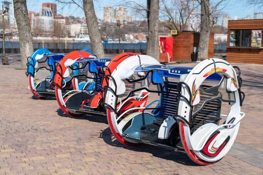 Colorful hover-like vehicles line the riverside walkway in a city, showcasing modern design. The area is sunny with trees and modern buildings in the background, indicating a vibrant setting.