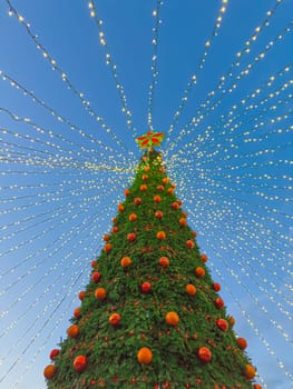 Colorful ornaments and bright lights adorn a large Christmas tree, creating a beautiful holiday display against a clear evening sky. The scene evokes a joyful festive spirit during the season.