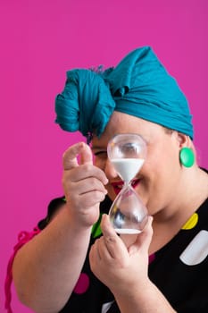 Portrait of a confident woman with a retro headwrap and colorful outfit, holding an hourglass in front of her face as a symbol of time and focus, with a vibrant pink background.