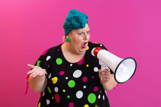 Confident woman in a colorful polka dot outfit and retro headwrap yelling into a megaphone, expressing strong emotion and attitude against a bold pink background.