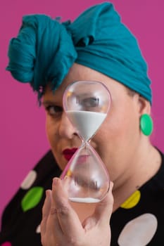 Portrait of a confident woman with a retro headwrap and colorful outfit, holding an hourglass in front of her face as a symbol of time and focus, with a vibrant pink background.