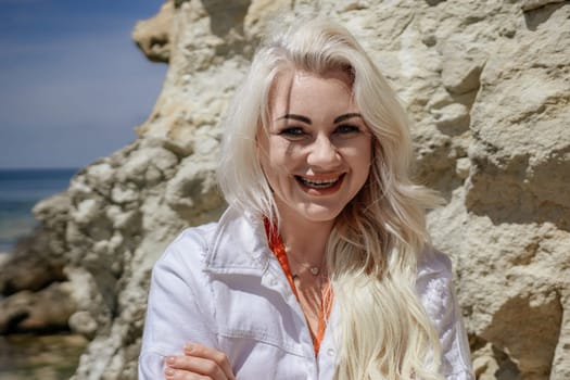 Blonde woman in white smiles on a sunny beach, rocky cliffs and ocean in the background. A happy and vibrant portrait.