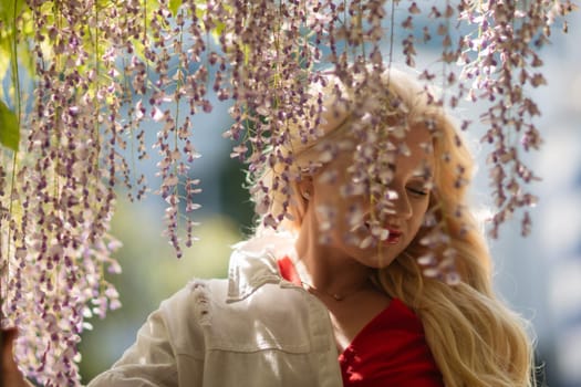 Blonde woman in red top stands under cascading wisteria. Natural light, soft focus. Relaxed pose in a garden setting.