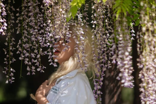 Blonde woman with a denim jacket standing beneath cascading wisteria blossoms, partially obscuring her face in a garden setting.