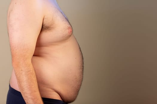 A man with a large belly is standing in front of a wall. The man's belly is protruding and he is wearing blue pants