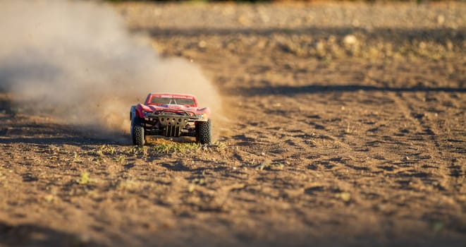 A red toy truck is driving through a dusty field. The truck is covered in dirt and the ground is covered in dust
