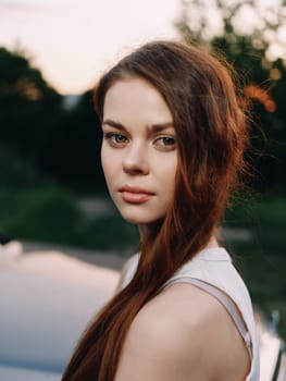 Beautiful young woman with flowing brown hair, wearing a stylish summer dress, looking contemplatively at the camera with a captivating sunset backdrop, showcasing tranquility and elegance.