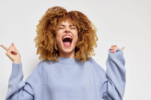 Joyful young woman with curly hair expressing happiness and excitement, wearing a comfortable blue sweater, posing against a clean white background, radiating positive energy and celebration