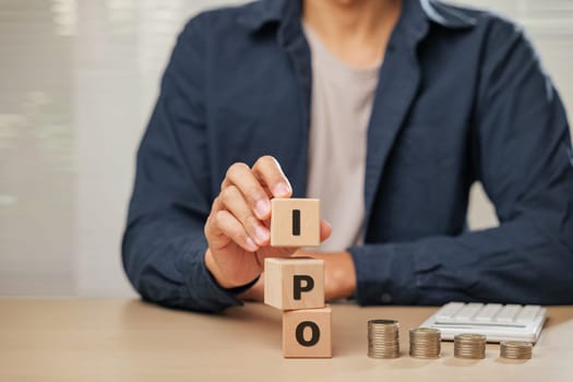 Close up businessman hand holding wooden cube blocks spelling IPO near coins and a calculator. Represents Initial Public Offering, planning, employee initiatives, and business financial accountability